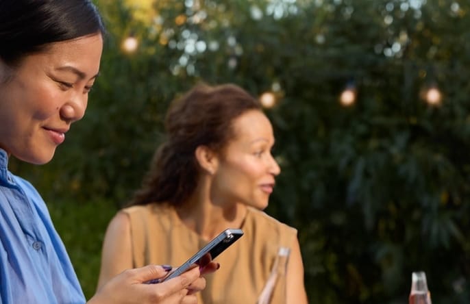 A woman in a blue blouse smiles while looking at her phone during an outdoor gathering with friends.