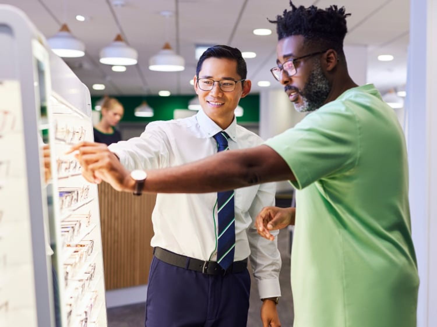 Two people wearing glasses examine a display of eyeglass frames together in a well-lit optical store.