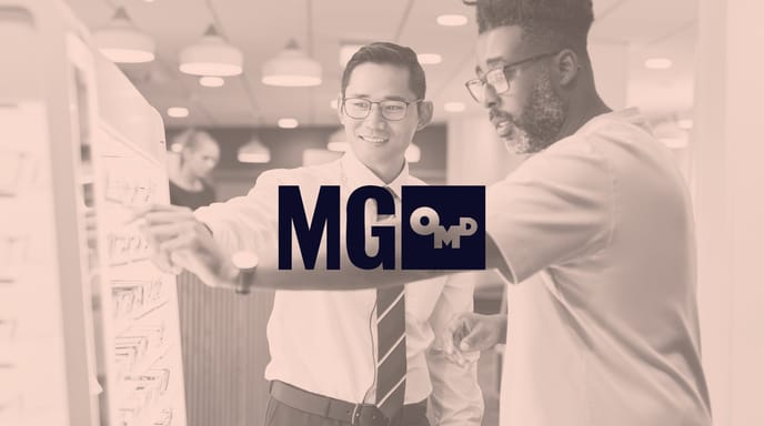 Two people wearing glasses examine a display of eyeglass frames together, shown in a sepia tone with the MG OMD logo centered over the image.