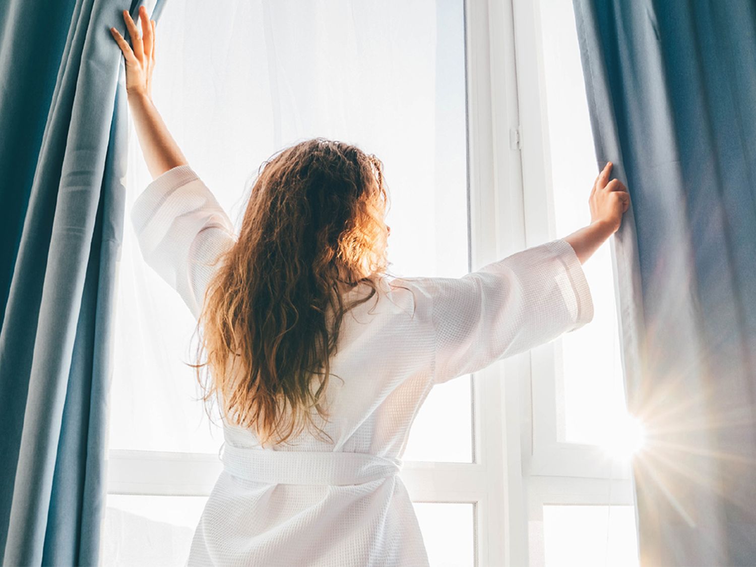 Woman wearing a white robe and opening window curtains to let the sun in