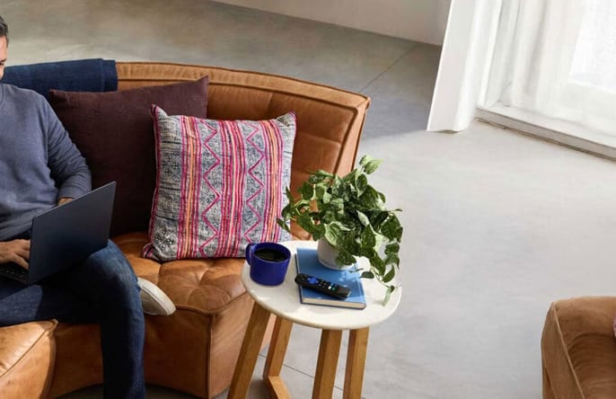 A man sits comfortably on a couch with colorful pillows, smiling while working on a laptop.