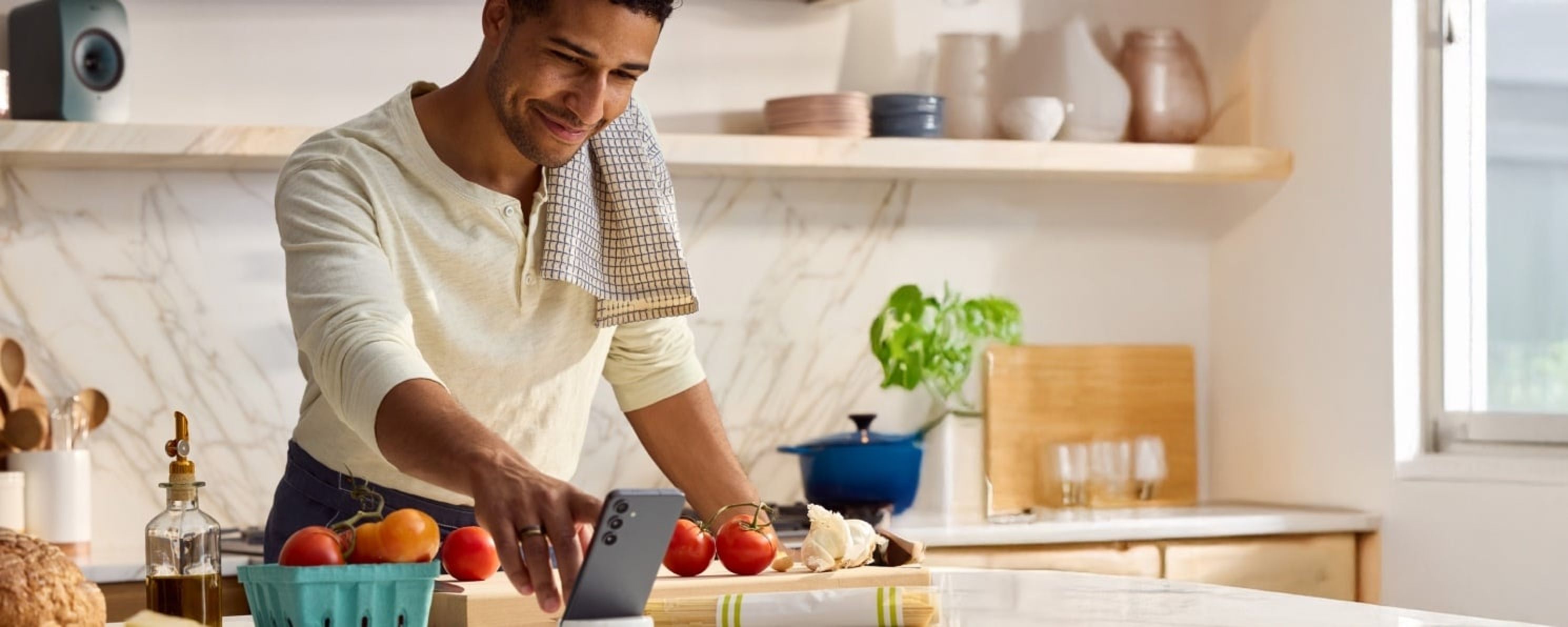 A man in a kitchen with a towel over his shoulder smiles while using his phone near fresh vegetables.
