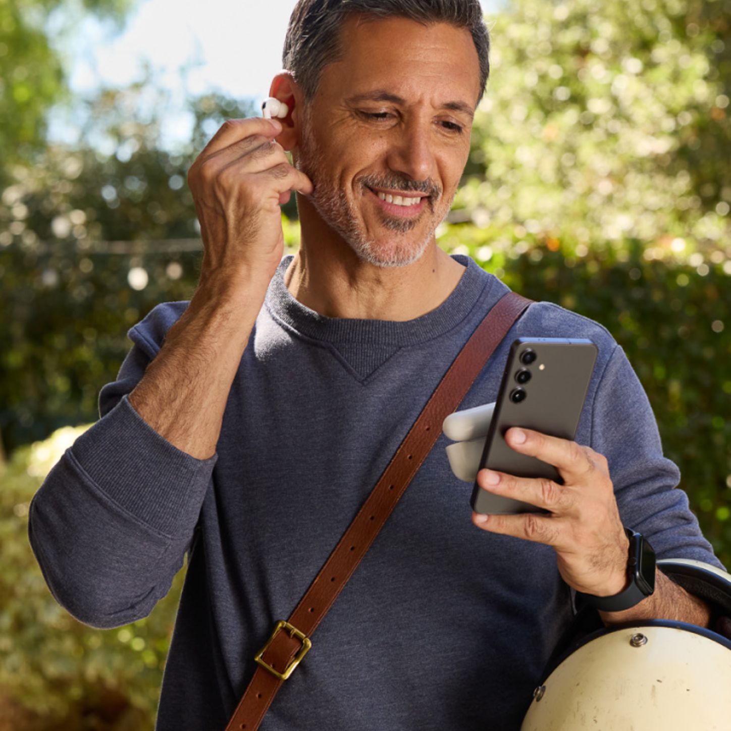 Un hombre sonriente lleva el smartphone en la mano y se coloca un auricular inalámbrico, lo cual sugiere que se dispone a escuchar contenido de audio mientras está fuera de casa.