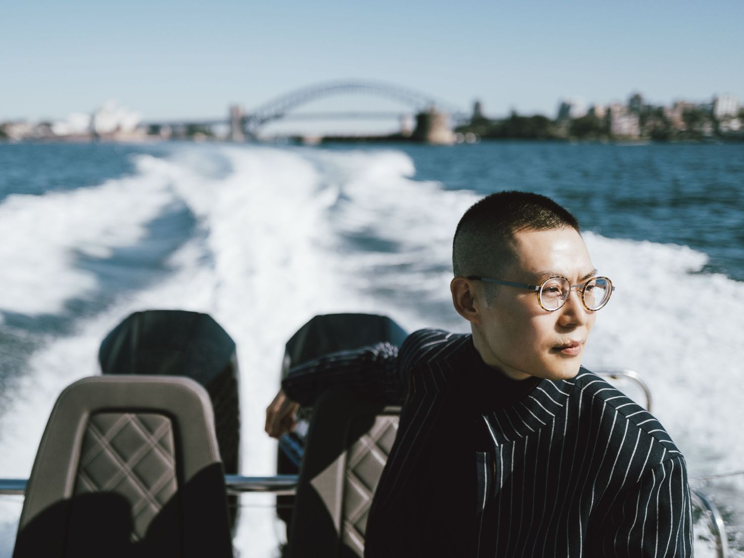 A stylish person wearing glasses and a pinstripe jacket rides on a speedboat with the Sydney Harbour Bridge and Opera House visible in the background.