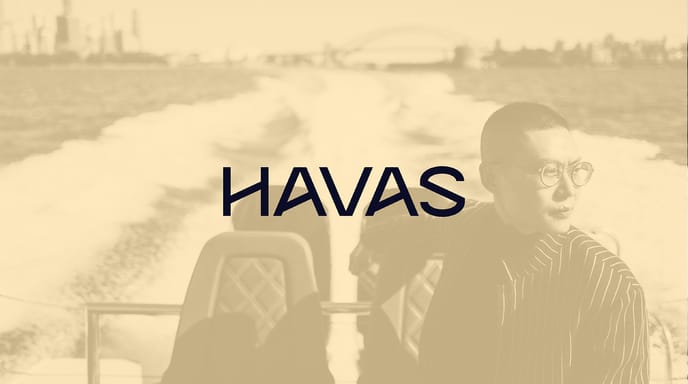 A stylish person wearing glasses and a pinstripe jacket rides on a speedboat with the Sydney Harbour Bridge in the distance, overlaid with a gold-toned filter and the HAVAS logo centered.