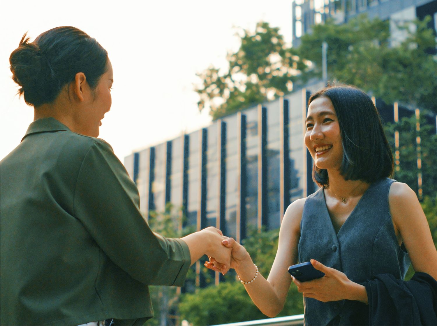 Two women smile and shake hands outdoors in a modern urban setting, one holding a phone and wearing a sleeveless outfit, with office buildings and greenery in the background.