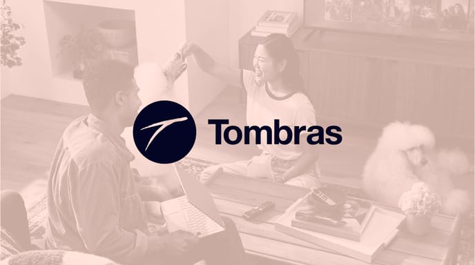 A man and woman smiling while interacting with a white poodle in a cozy living room, overlaid with the Tombras logo.