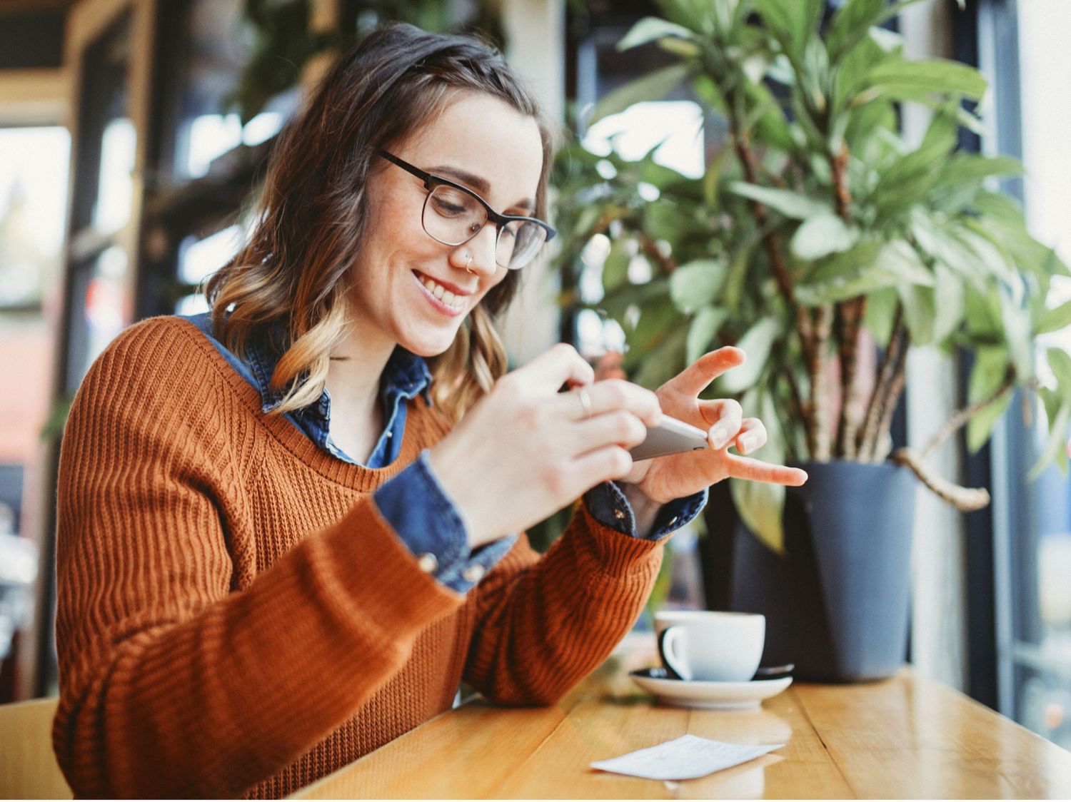 A smiling woman wearing glasses and a rust-colored sweater takes a photo of a receipt with her smartphone in a cozy café.