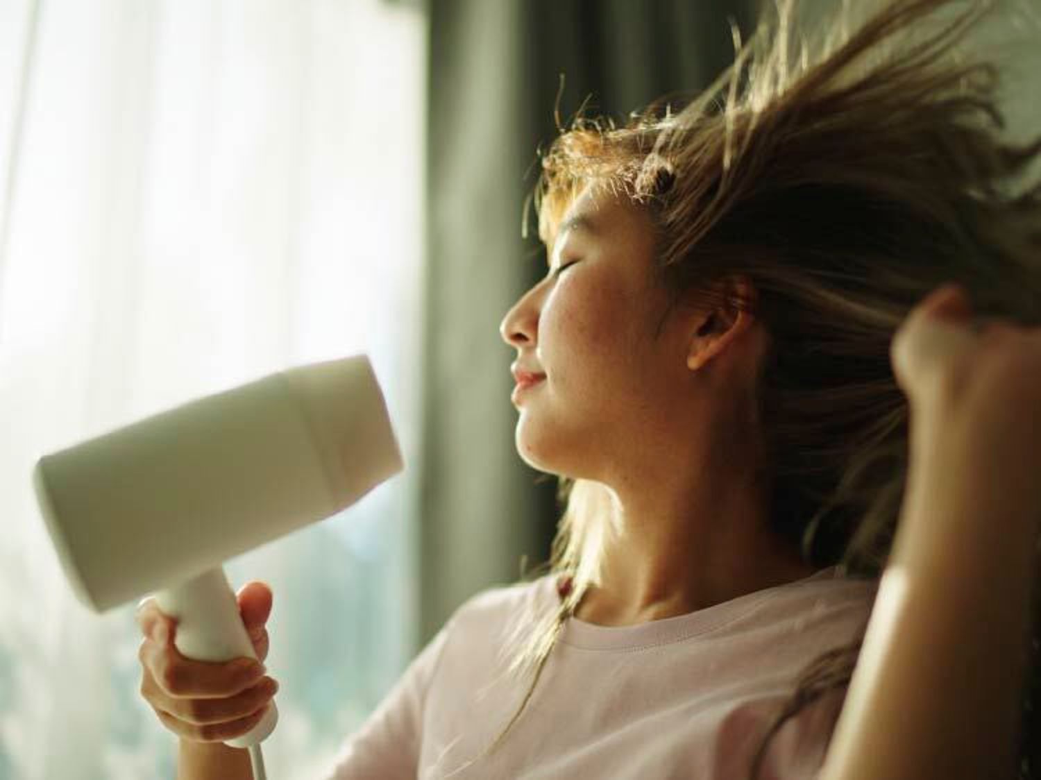 A woman with closed eyes enjoys drying her hair with a white hair dryer as sunlight streams through a nearby window.