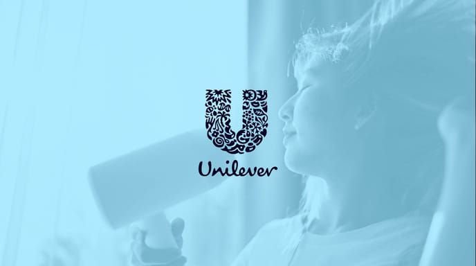 A woman enjoys drying her hair with a white hair dryer in soft natural light, overlaid with the Unilever logo on a blue-tinted background.