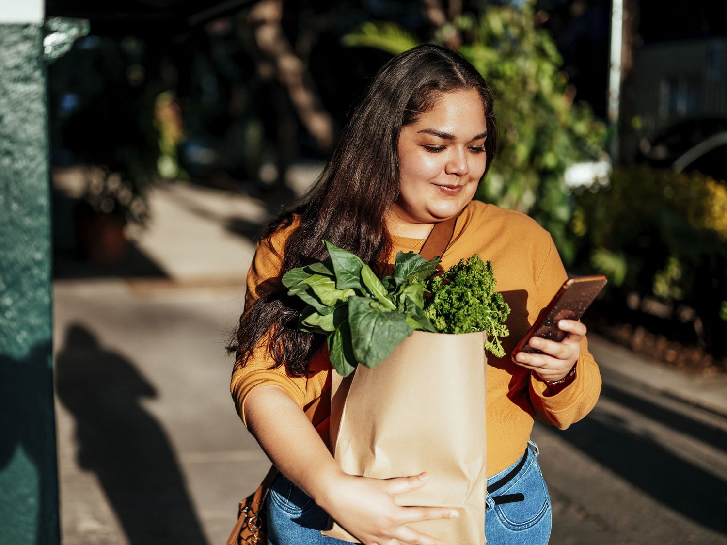 A woman holding a paper bag full of leafy greens smiles while looking at her smartphone outdoors on a sunny day.