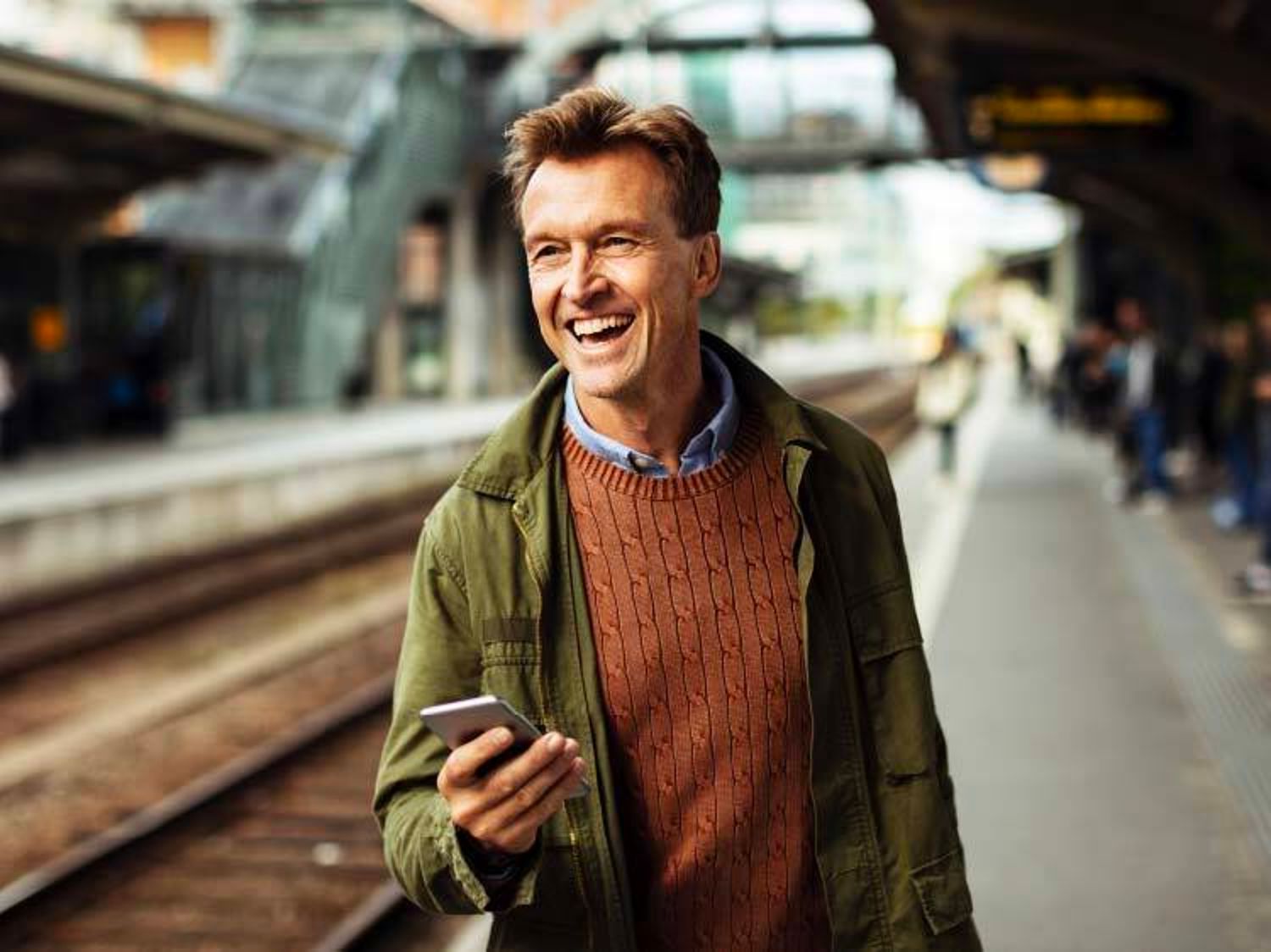 A smiling man holding a smartphone stands on a train platform, wearing a green jacket over a brown cable-knit sweater.