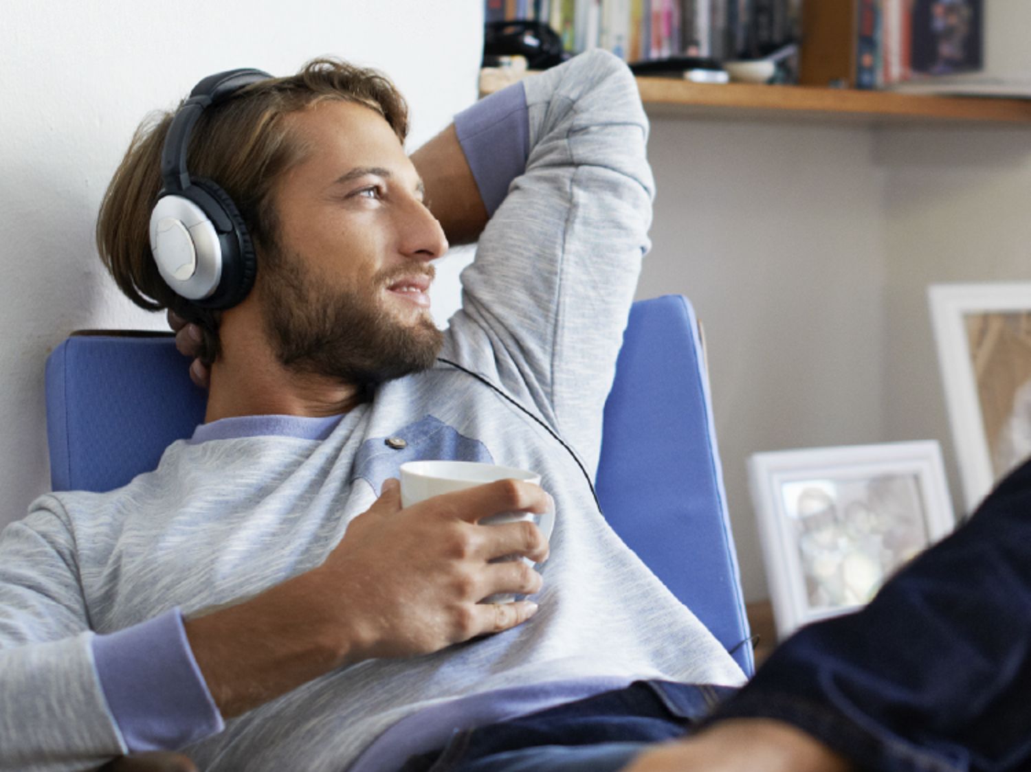A man relaxing on a chair, wearing headphones and holding a cup, with a content expression.