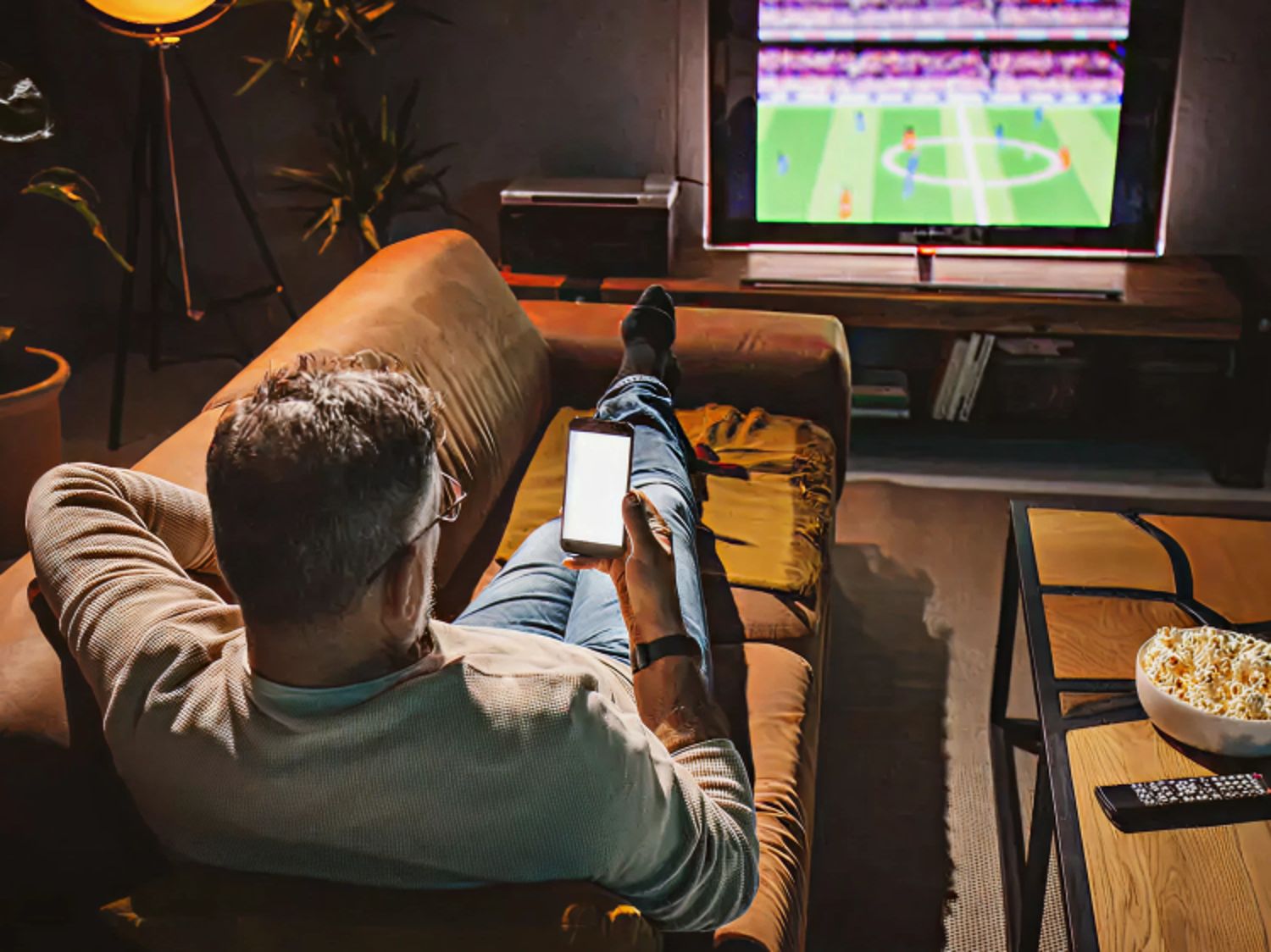 A man relaxes on a couch while watching a soccer game on TV and browsing his smartphone.