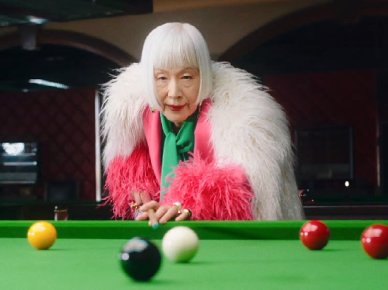 鮮やかな毛皮のコートを着た白髪ボブのスタイリッシュな年配の女性。色とりどりのボールが並ぶビリヤード台に身を乗り出して、ショットを打とうとしている。