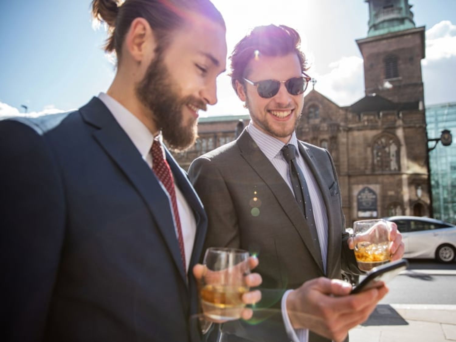 晴れた日の屋外で、飲み物を片手に笑顔でスマホを眺めているスーツ姿の2人の男性