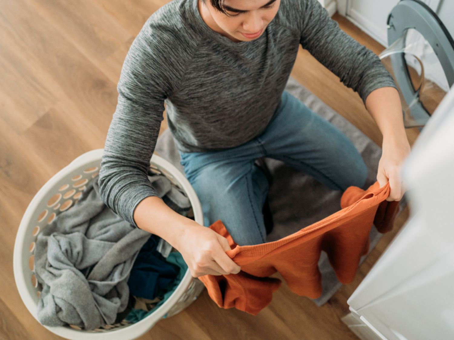 Person sorting laundry, holding an orange shirt above a laundry basket filled with clothes next to a washing machine.