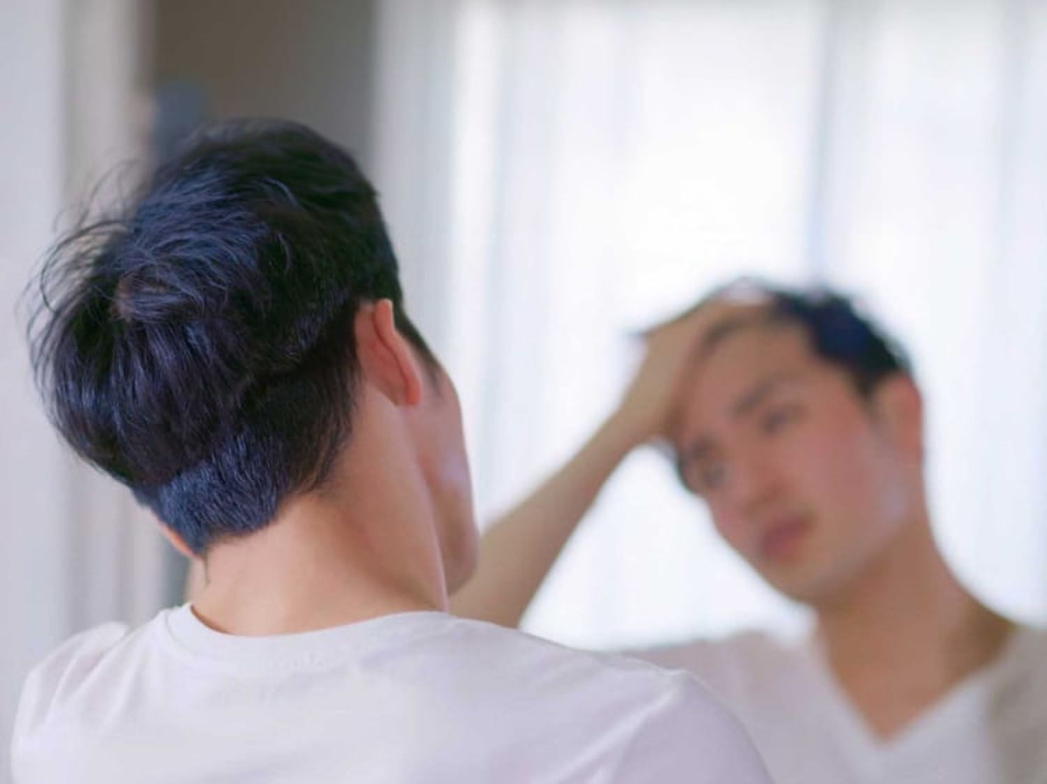 柔らかな光が差し込む部屋で、鏡に映る自分の姿を見ながら手ぐしで髪を整える男性。