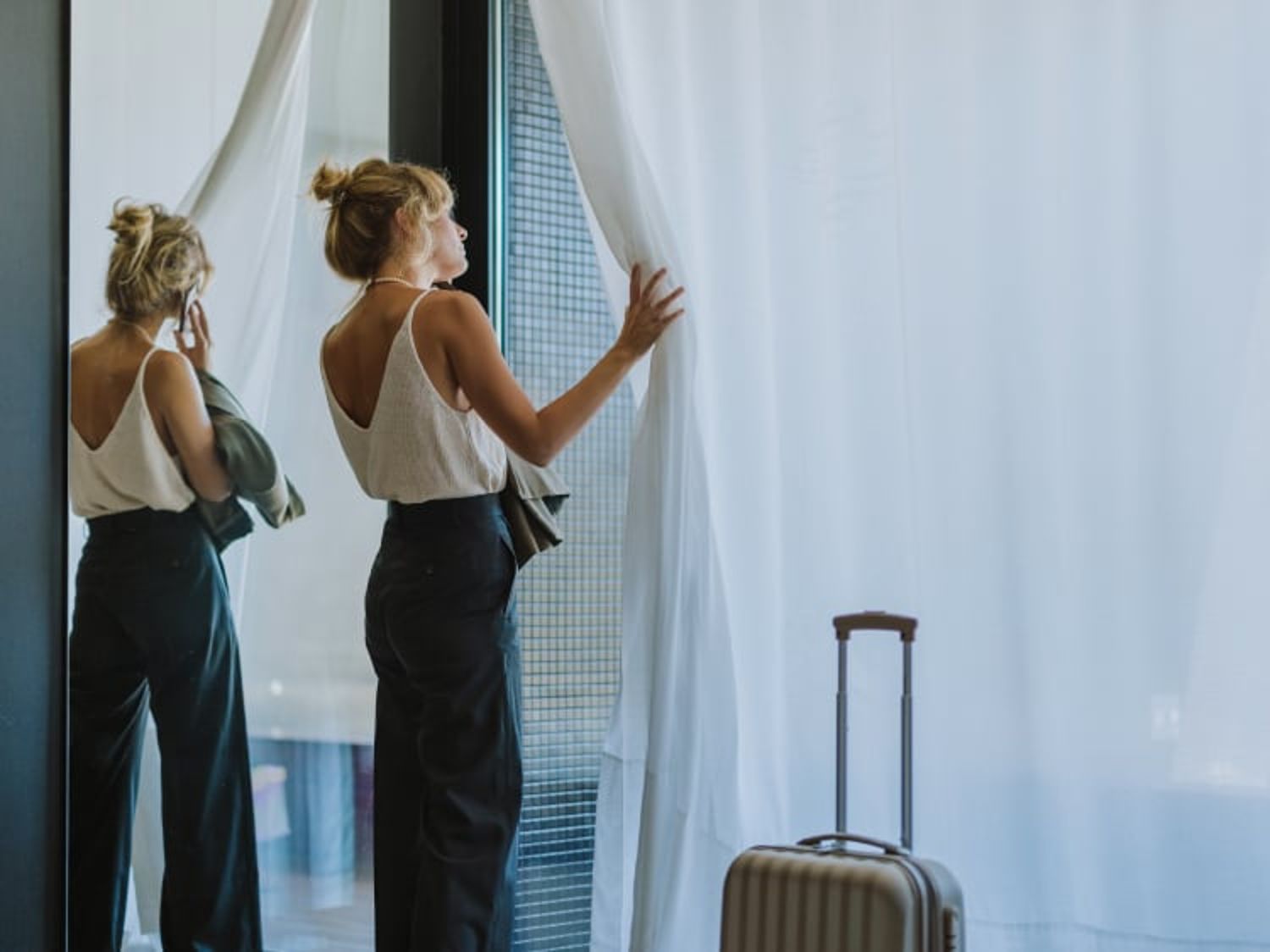 Traveler standing near a suitcase and looking out of a large window with sheer curtains.