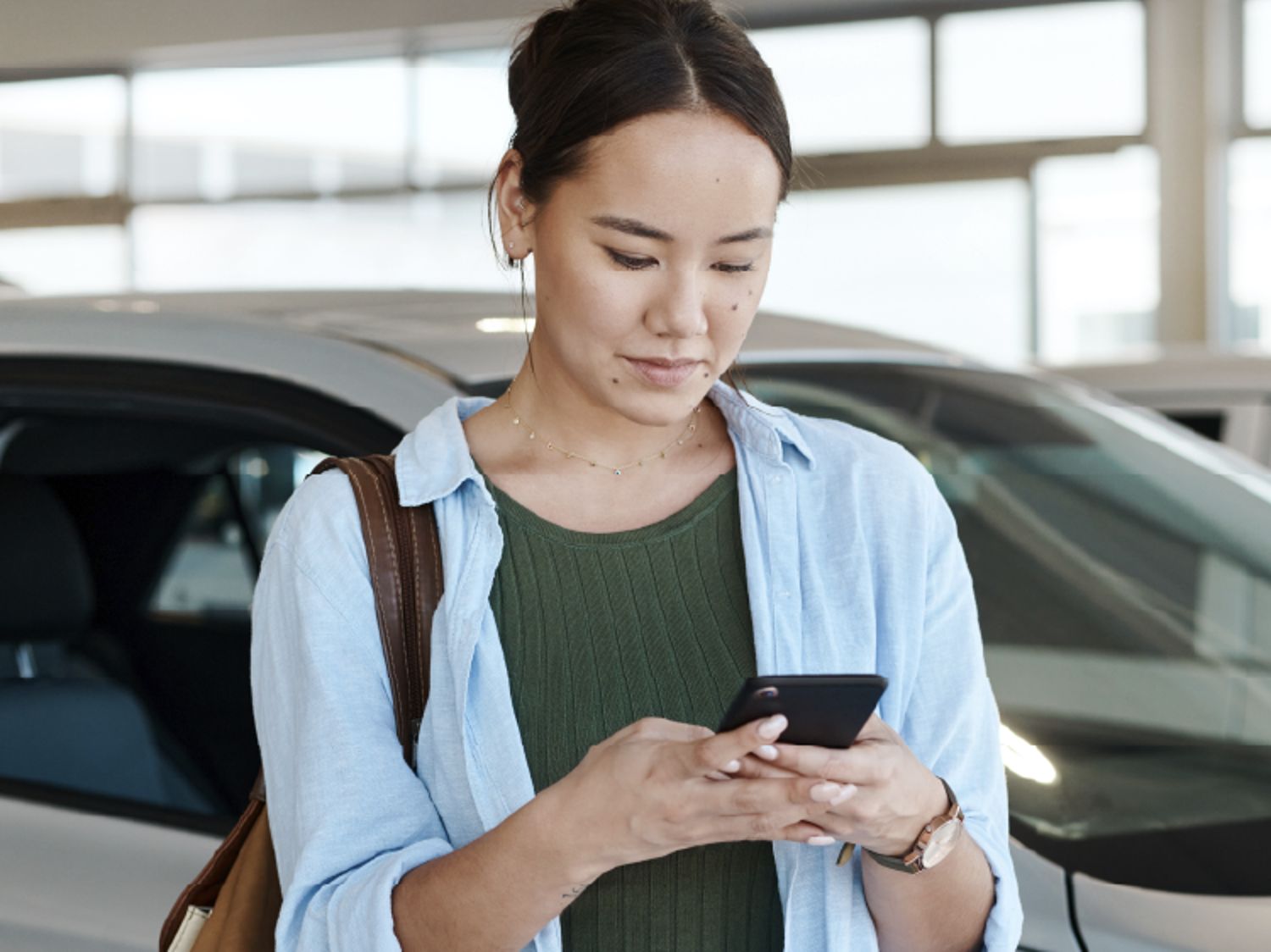 A woman stands next to a car in a showroom, focused on her phone while wearing a green top, blue shirt, and carrying a brown bag.