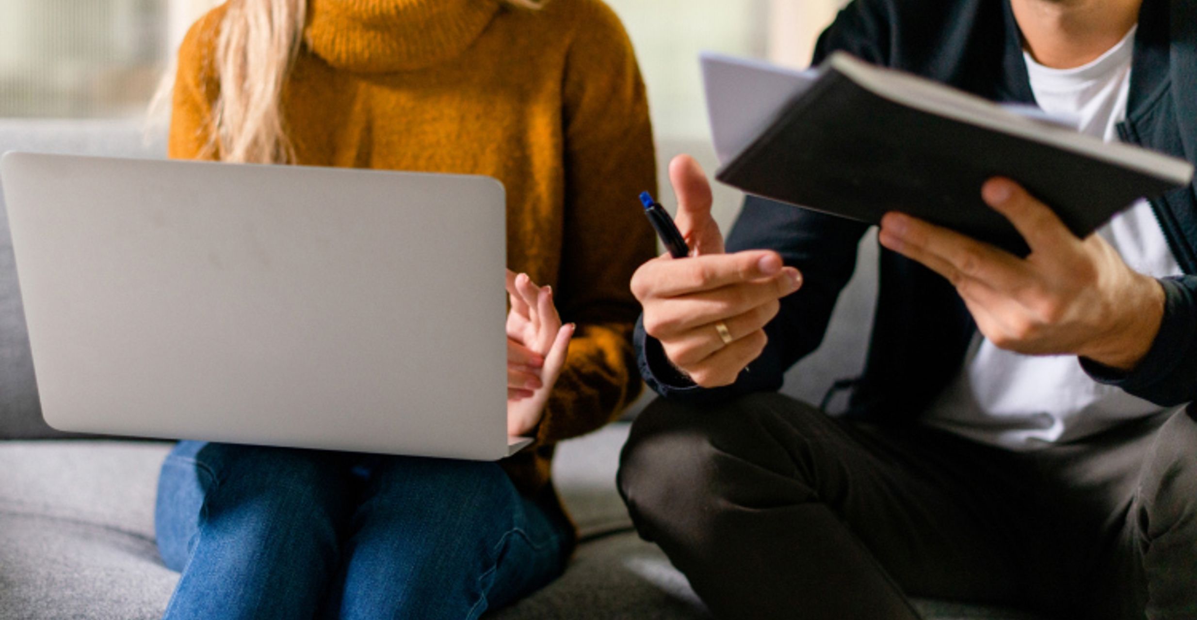 Two people sitting on a couch working together, one using a laptop and the other holding a notebook and pen.