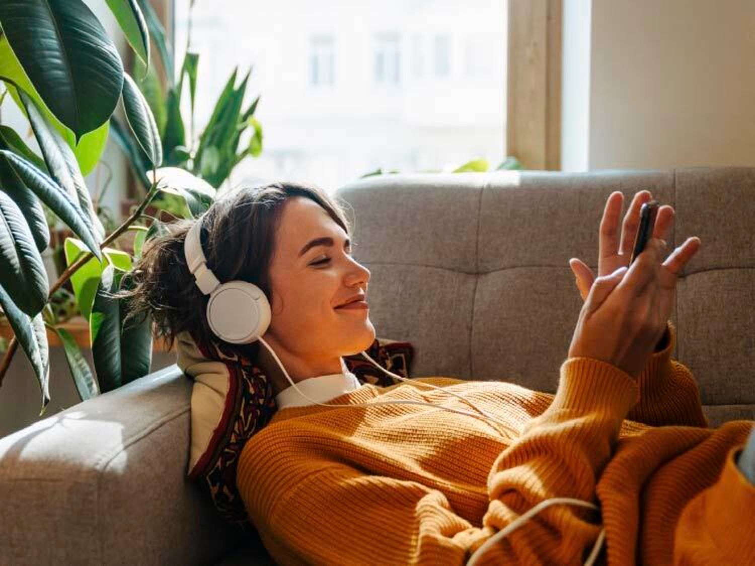 A woman relaxes on a couch wearing headphones and smiling while looking at her phone.