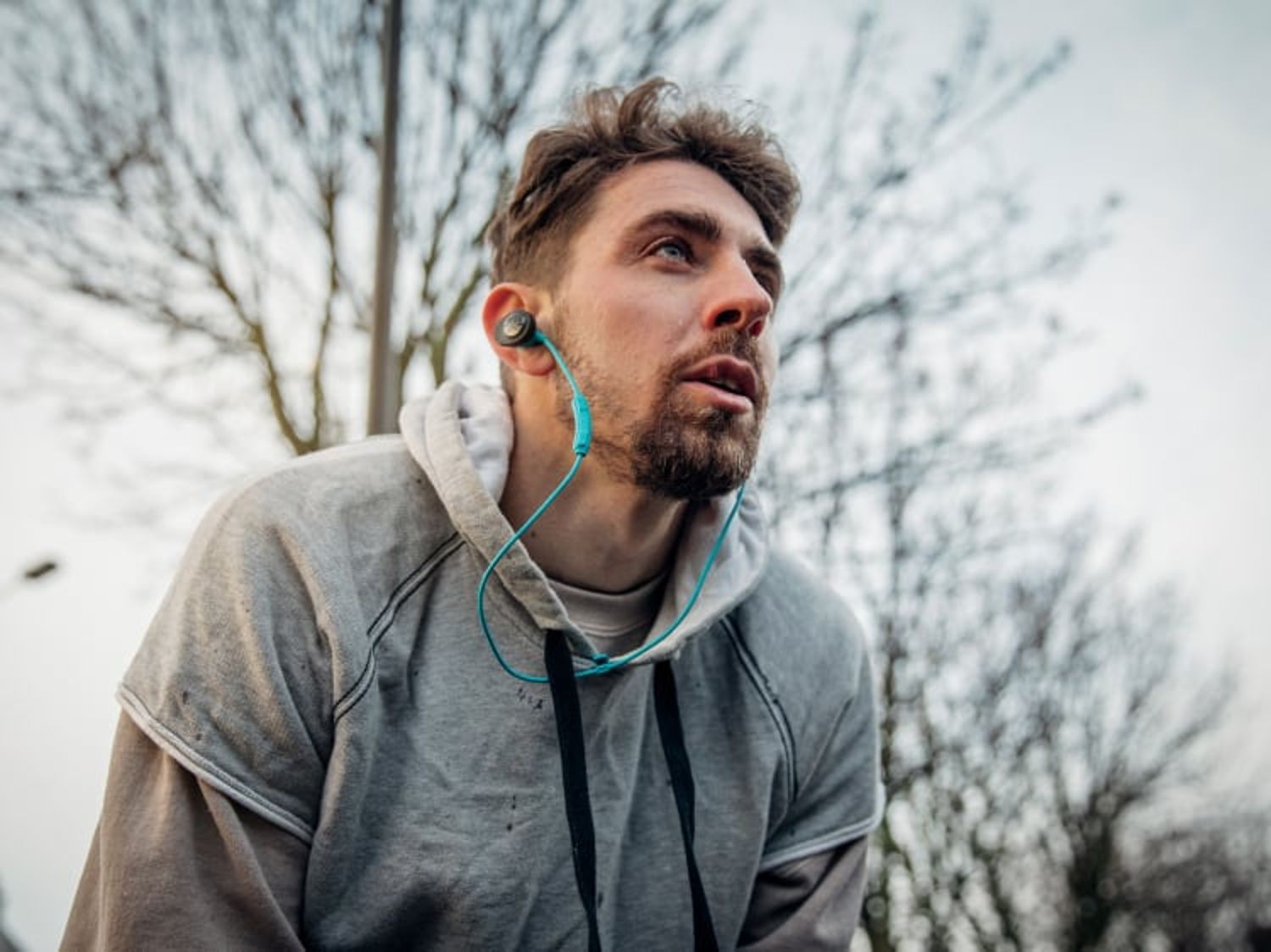 A man wearing a hoodie and wired earbuds pauses to catch his breath during an outdoor workout on a chilly day.