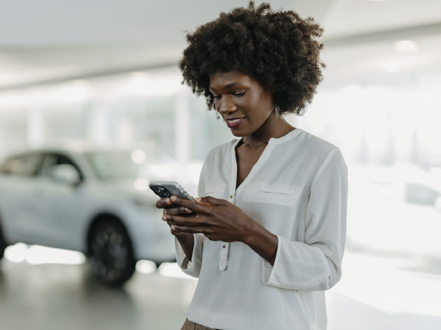 A woman in a white blouse smiles while using her smartphone inside a bright, modern showroom with a white car in the background.