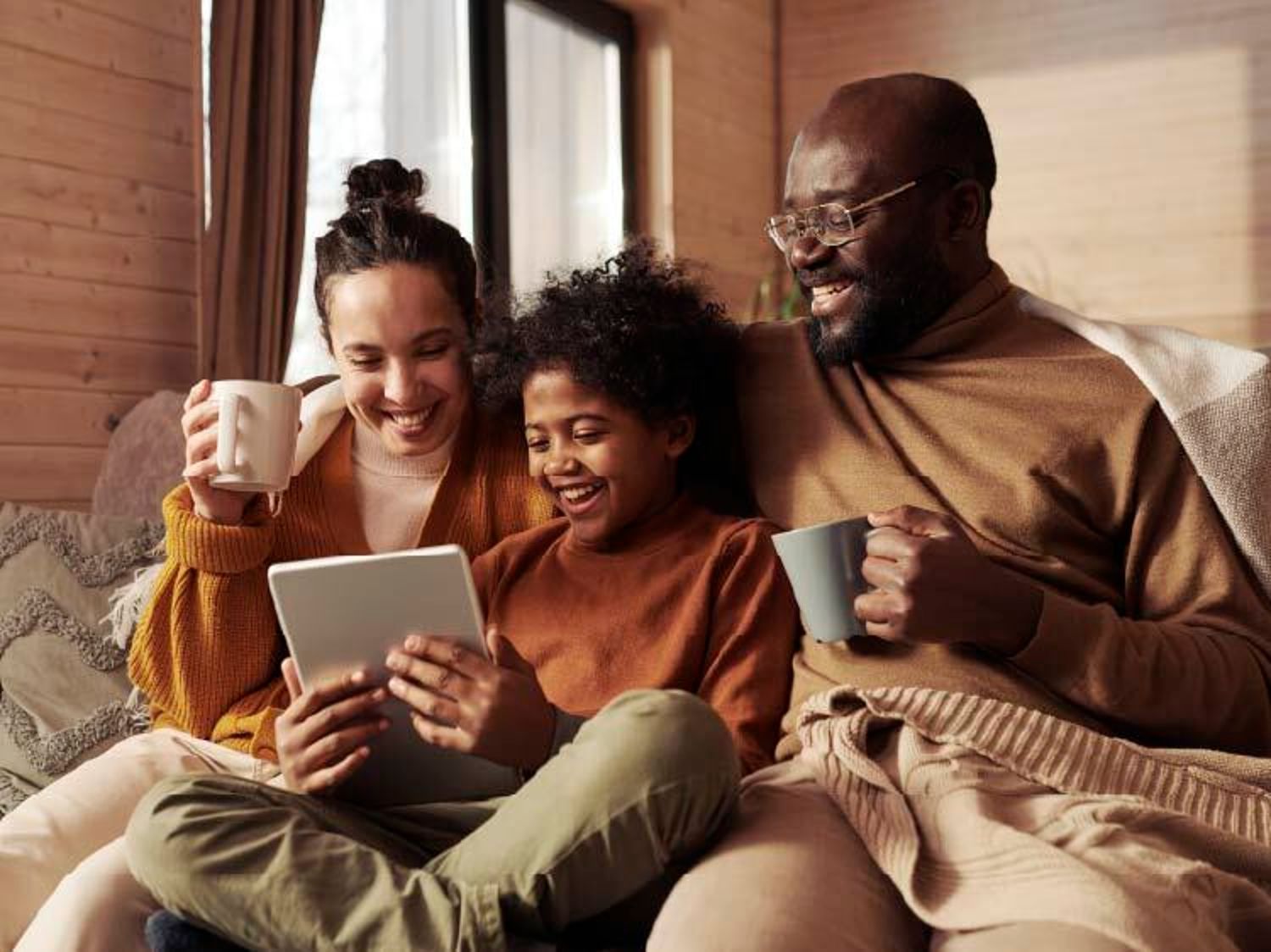 A mom, dad and child all holding each other and smiling while drinking coffees and looking at a tablet.