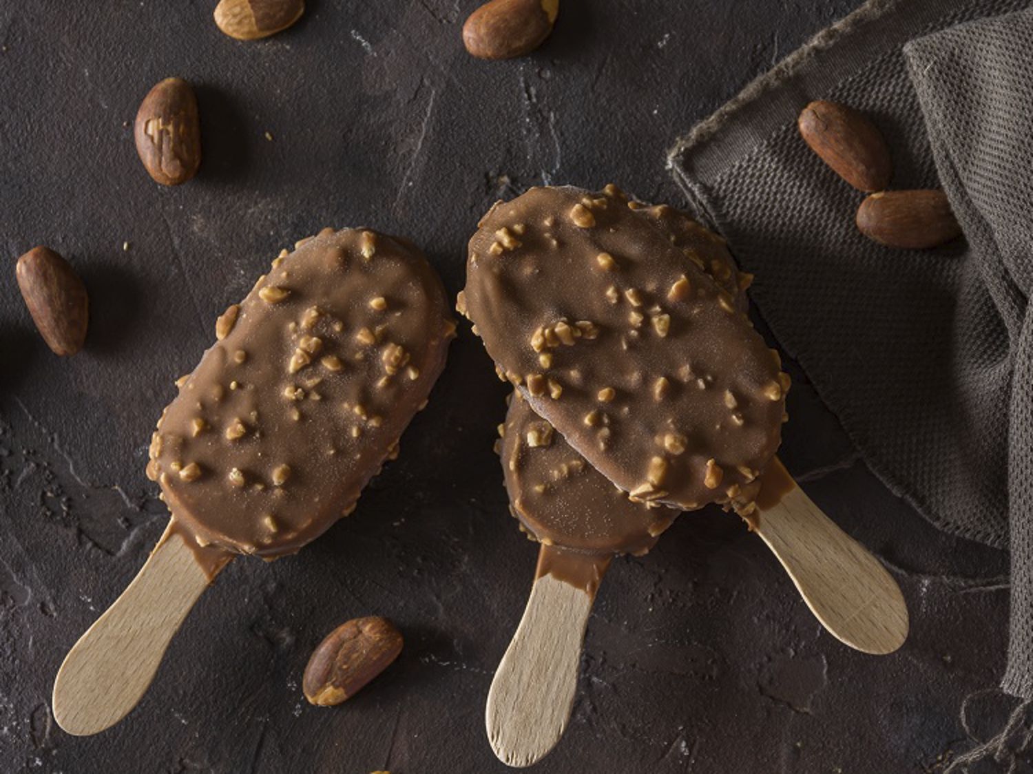 Two chocolate-coated ice cream bars with nuts on wooden sticks, placed on a dark textured surface with scattered cocoa beans.