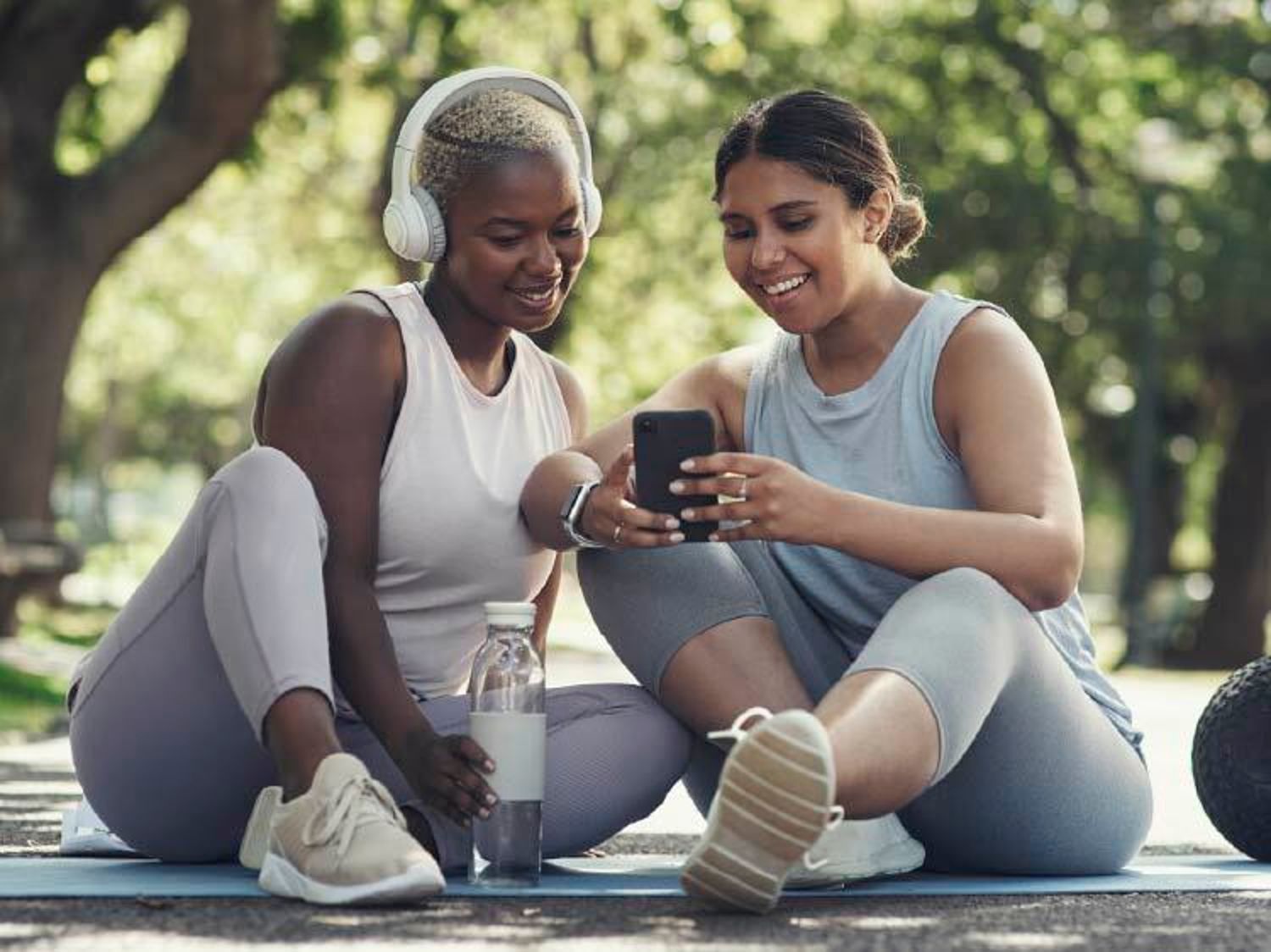Two women in athletic wear sit outdoors, smiling while looking at a smartphone, with one wearing headphones and the other holding the phone.