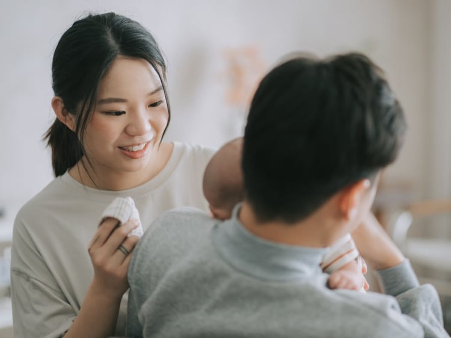 A smiling woman looks at a man holding a baby on his shoulder, as she gently holds a cloth in a softly lit, cozy setting.