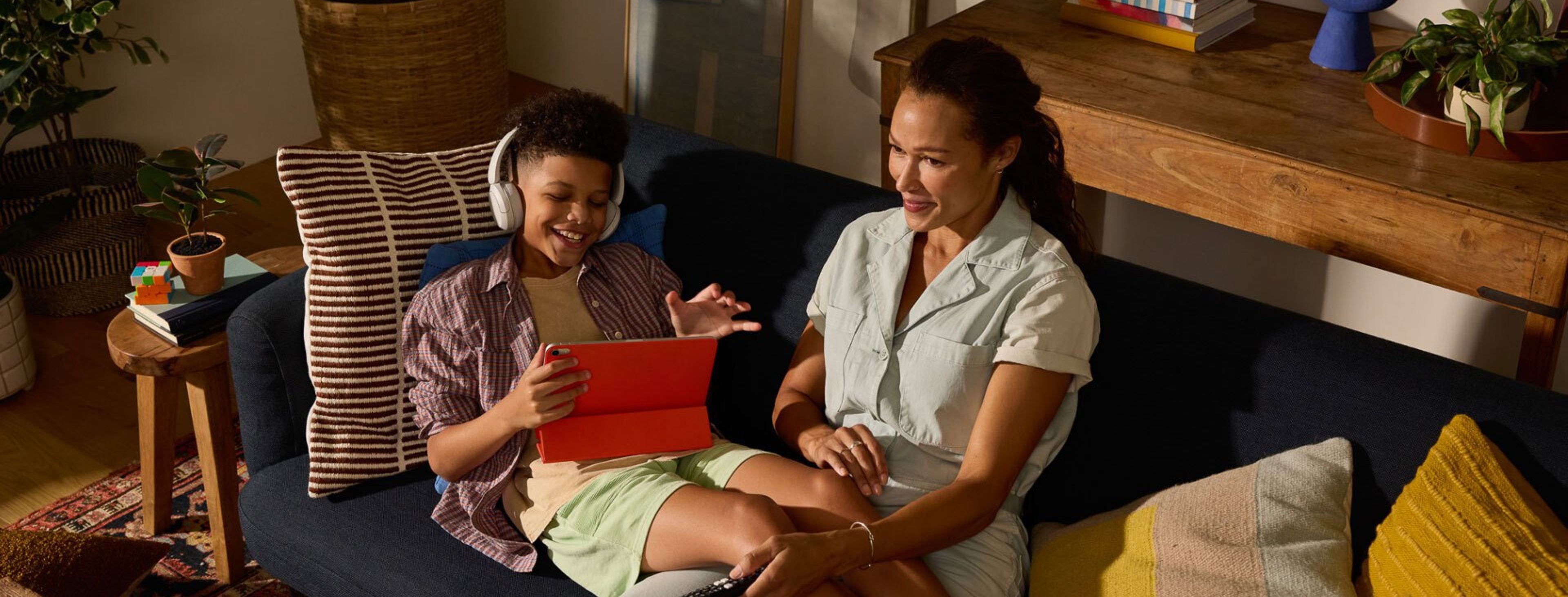 A woman and child sit on a cozy couch in a warmly lit living room, smiling and enjoying digital content together on a tablet, with the child wearing headphones.
