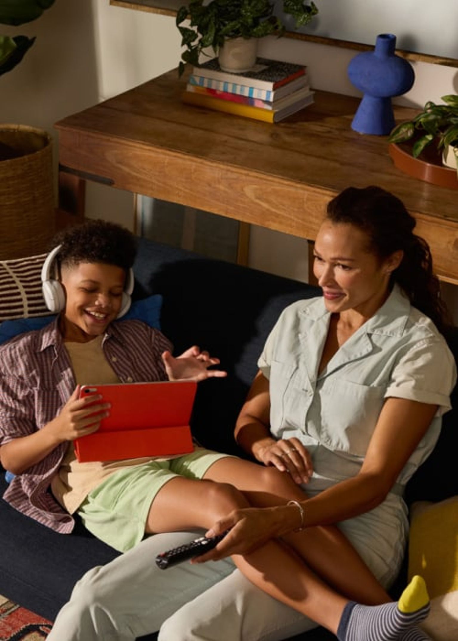 A woman and child sit on a cozy couch in a warmly lit living room, smiling and enjoying digital content together on a tablet, with the child wearing headphones.