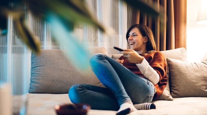 A woman relaxes on a couch, smiling while watching TV with a remote.