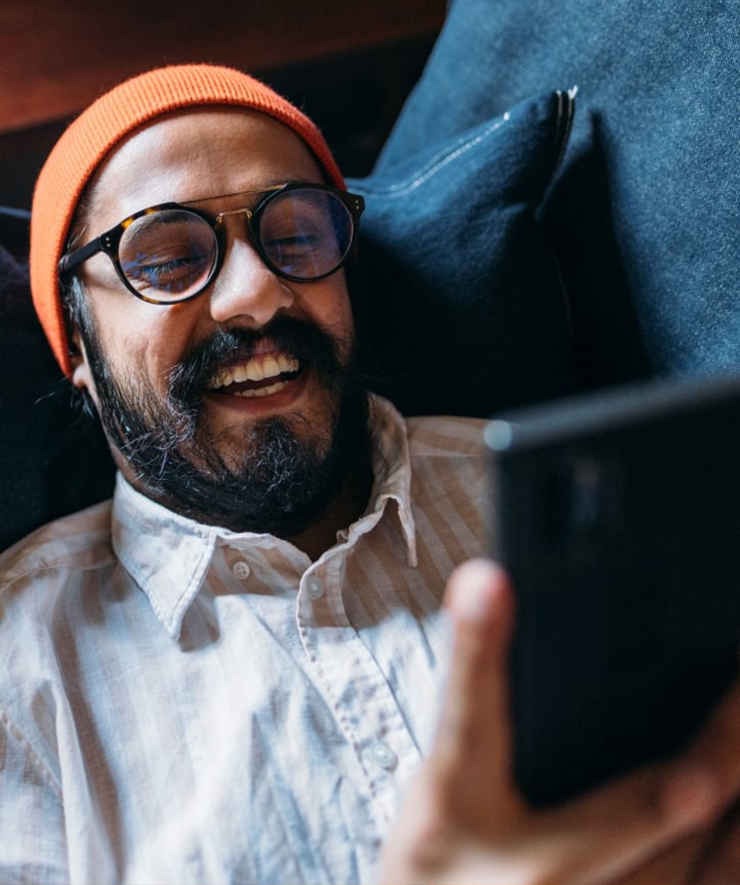 Smiling man in a beanie and glasses lounging on a couch, enjoying content on his smartphone