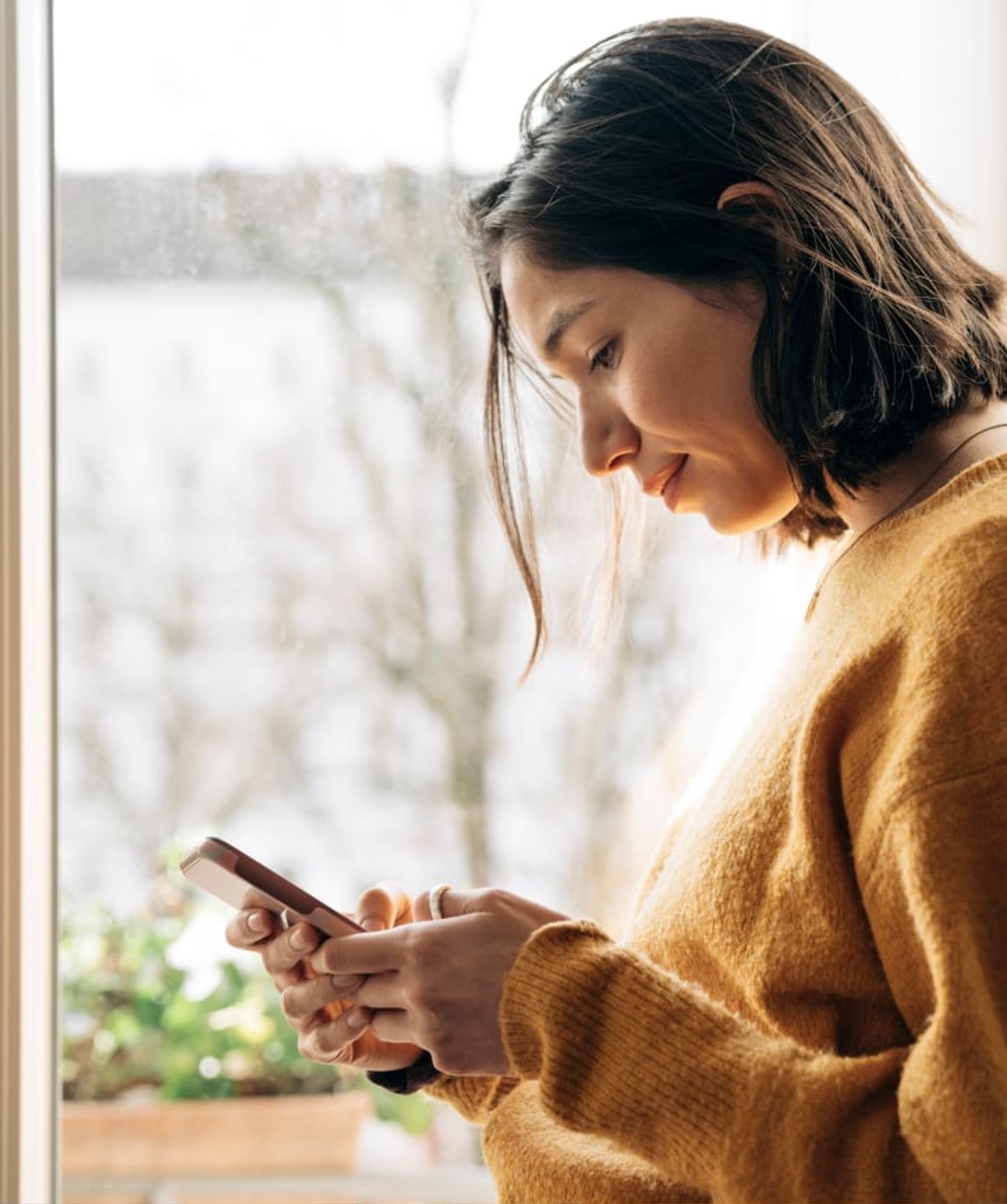 Young woman in a cozy sweater looking at her smartphone near a window on a winter day