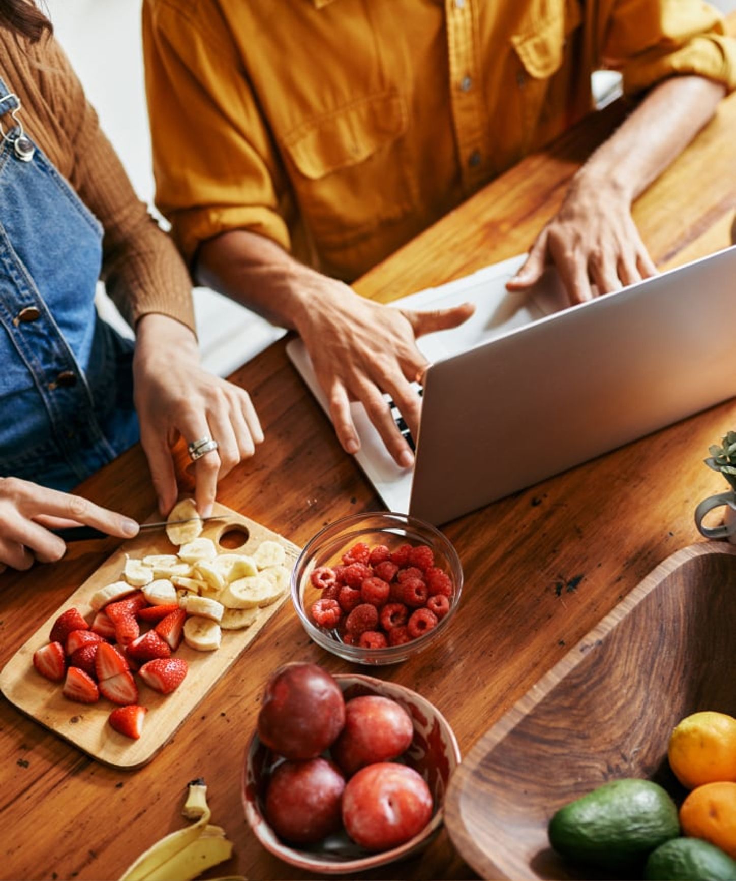 Close-up of a couple preparing fresh fruit while working on a laptop at a wooden kitchen table