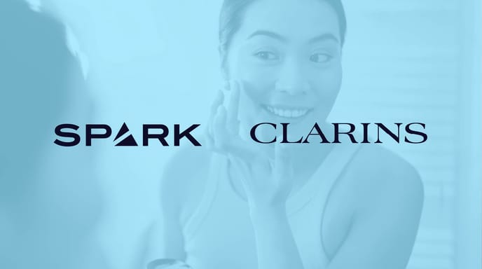 Woman looking in the mirror and smiling, while doing her skincare, with a blue overlay over and Spark + Clarins logos over the top.