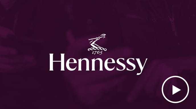 Hennessy logo with a play button overlay, set against a dark purple background with two people holding phones faintly visible.