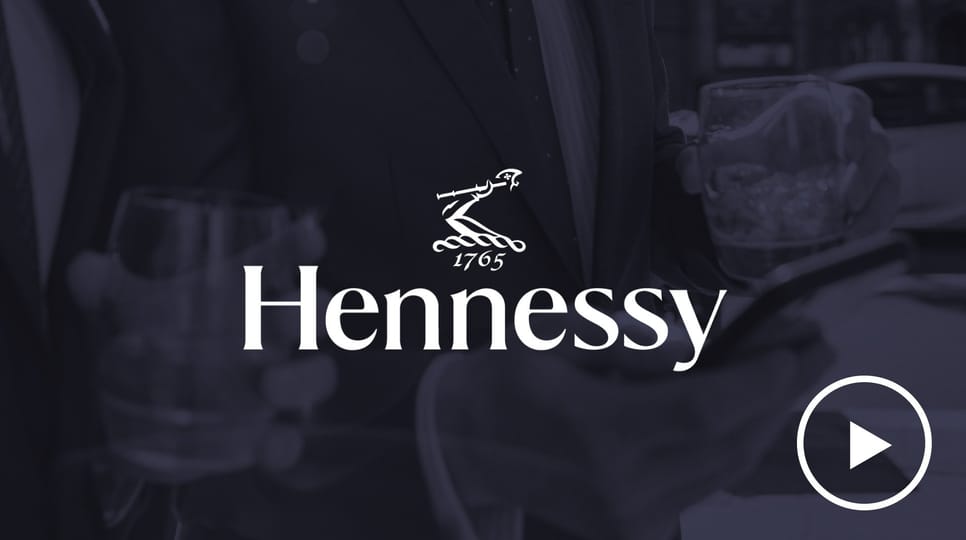 Hennessyのグラスを持ち、スマートフォンを見ているスーツ姿の2人の男性。その上にHennessyのロゴと再生ボタンが重ねられている。
