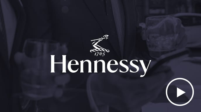Hennessyのグラスを持ち、スマートフォンを見ているスーツ姿の2人の男性。その上にHennessyのロゴと再生ボタンが重ねられている。