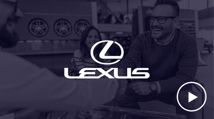 A cheerful man in a red sweater shakes hands across a table while a smiling woman looks on, overlaid with the Lexus logo and a play button in a monochrome showroom setting.