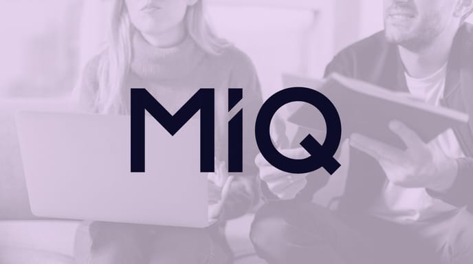Logo for MiQ displayed over a purple-tinted background of two people working on a laptop and notebook.