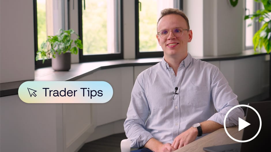 A smiling person wearing glasses and a light blue shirt sits in a bright modern office with plants and large windows, with a “Trader Tips” graphic and a play button icon on screen.