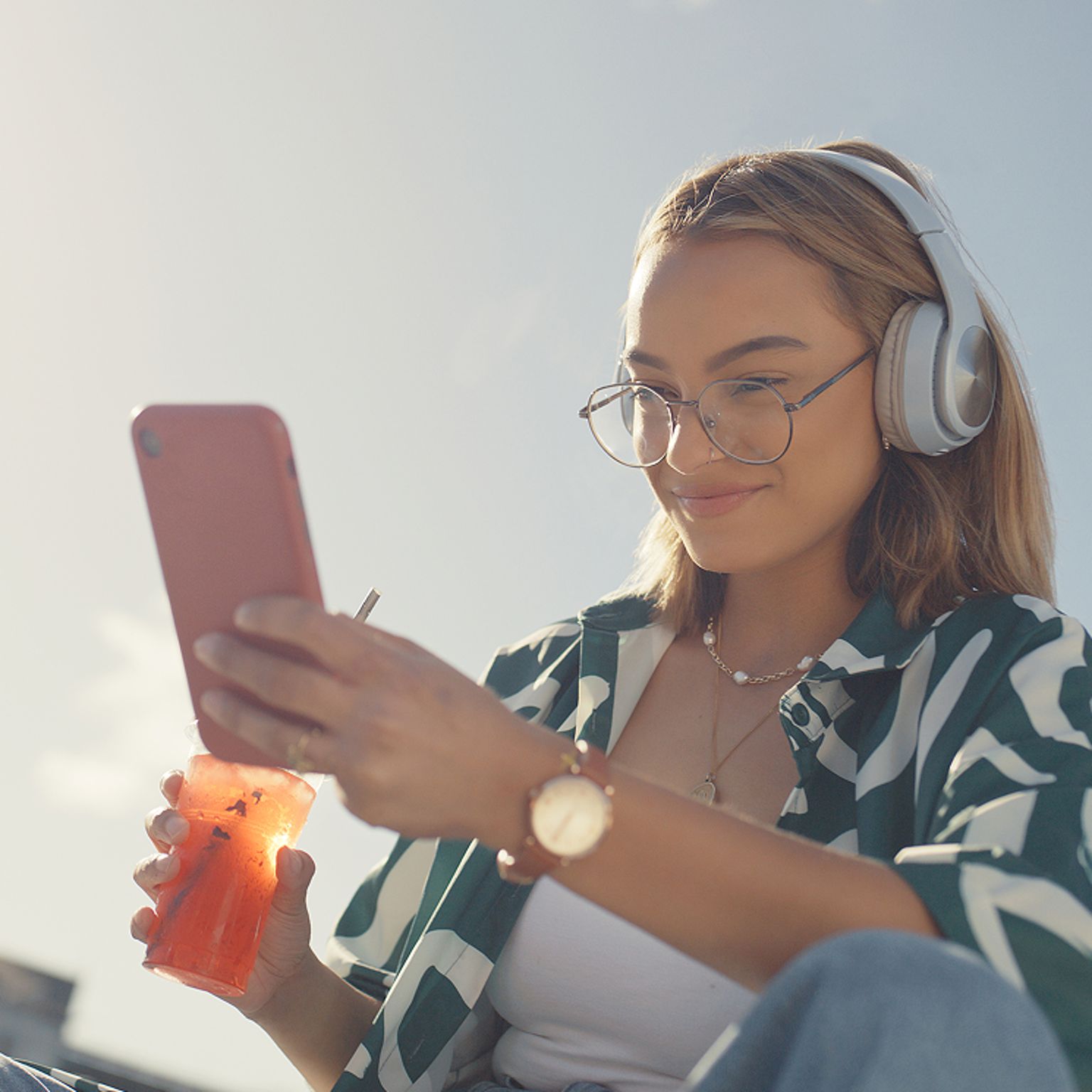 Woman wearing headphones using smartphone outdoors while enjoying a drink