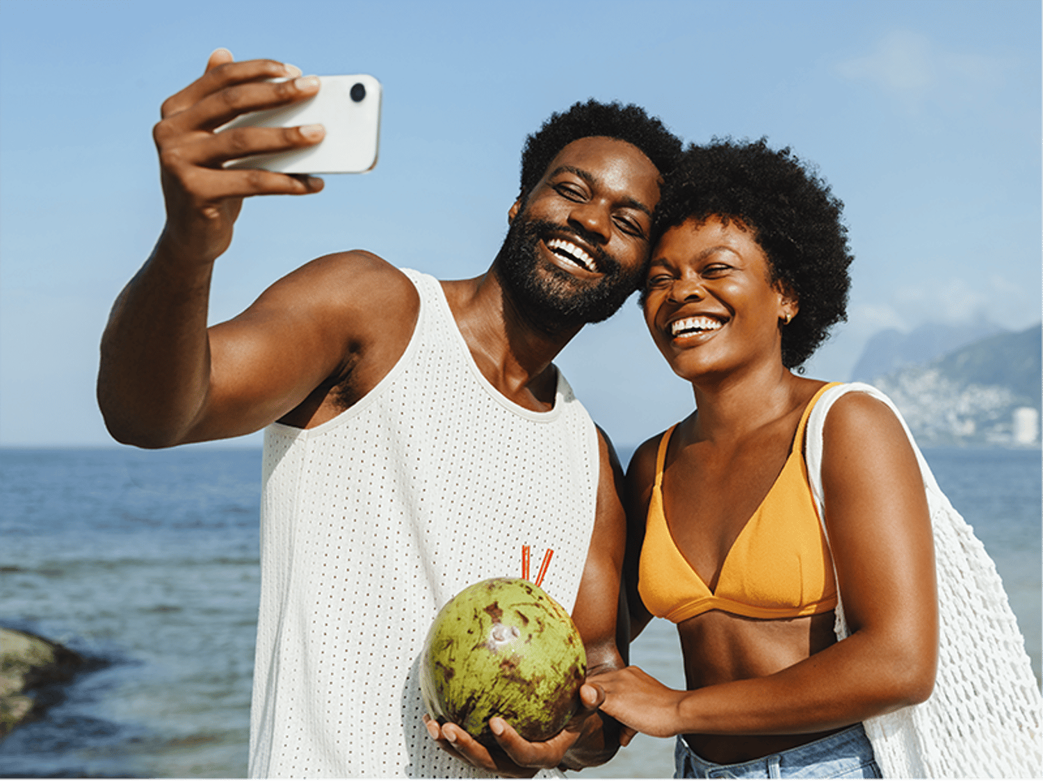 A happy couple taking a selfie on the beach while holding a fresh coconut with straws.
