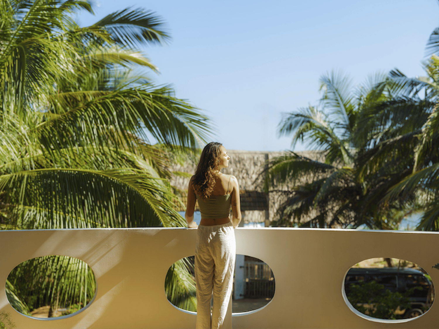 Image shows a woman standing on a balcony overlooking palm trees and basking in the sun