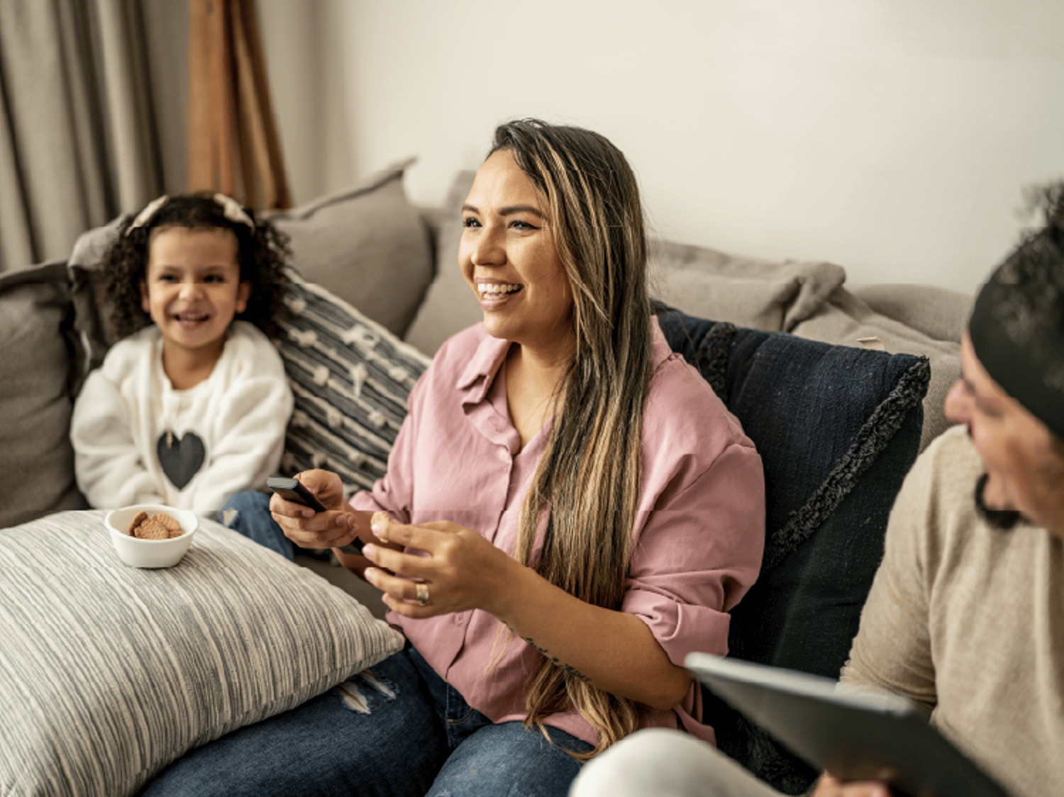 A cheerful family lounges on a couch sharing snacks and conversation, with a young girl smiling brightly beside a woman holding a remote and cookies.