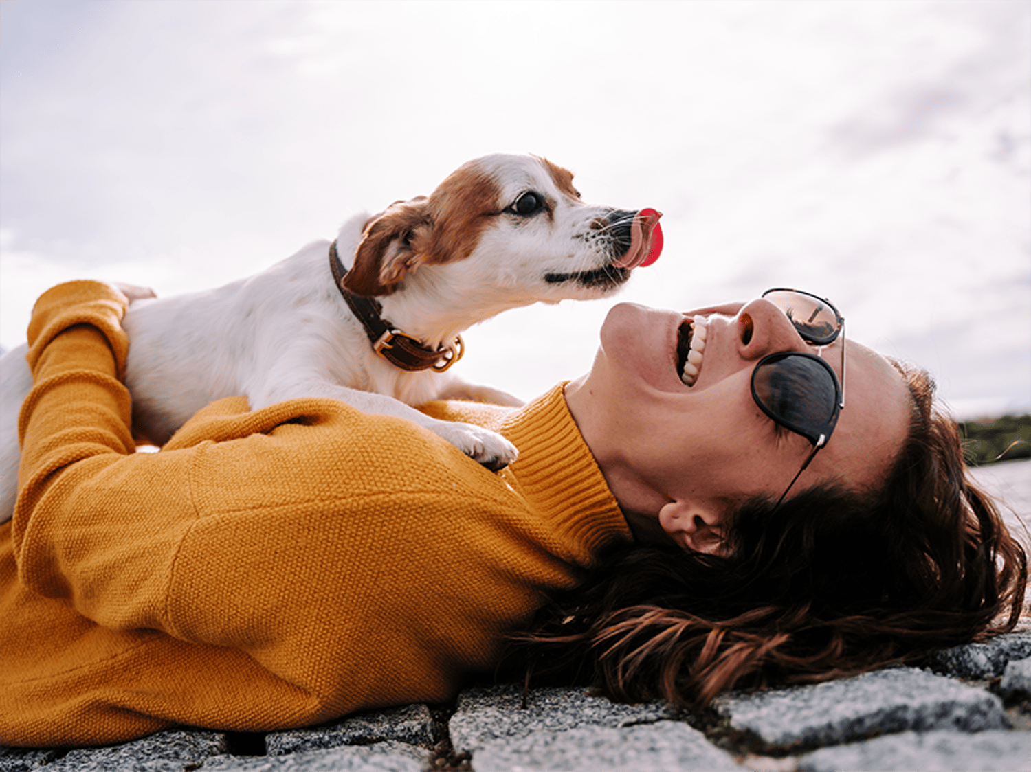 A joyful woman in a sweater and sunglasses lying on the ground, laughing as a small dog playfully licks its nose above her.